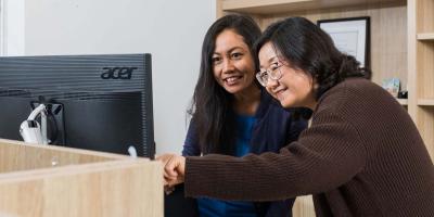 Two women in front of a computer screen, smiling, one woman is pointing to the computer screen